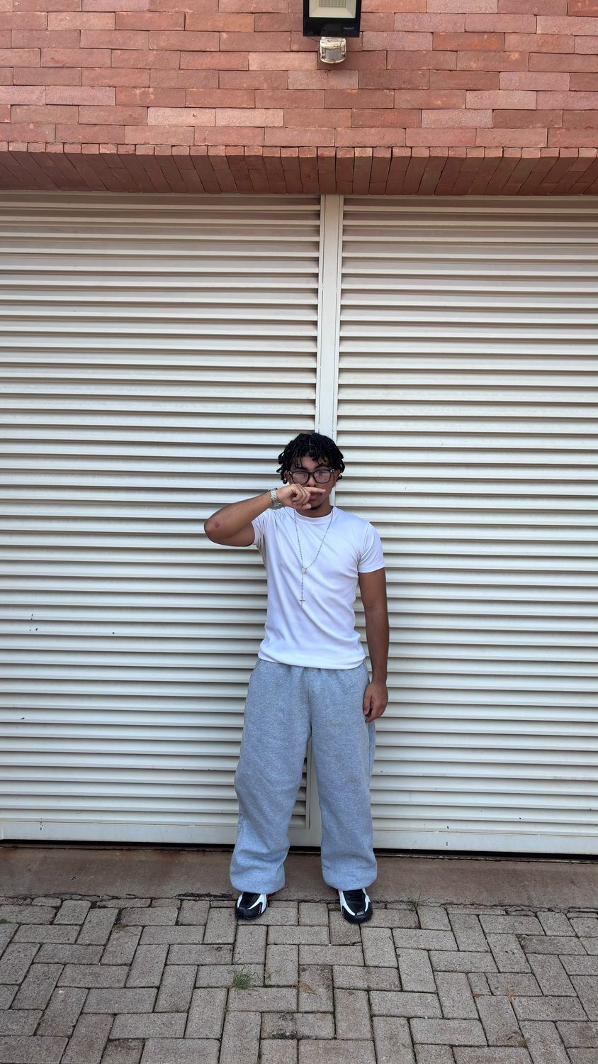 Feliphe de pé diante do portão branco, mão cobrindo o rosto, calça de moletom cinza